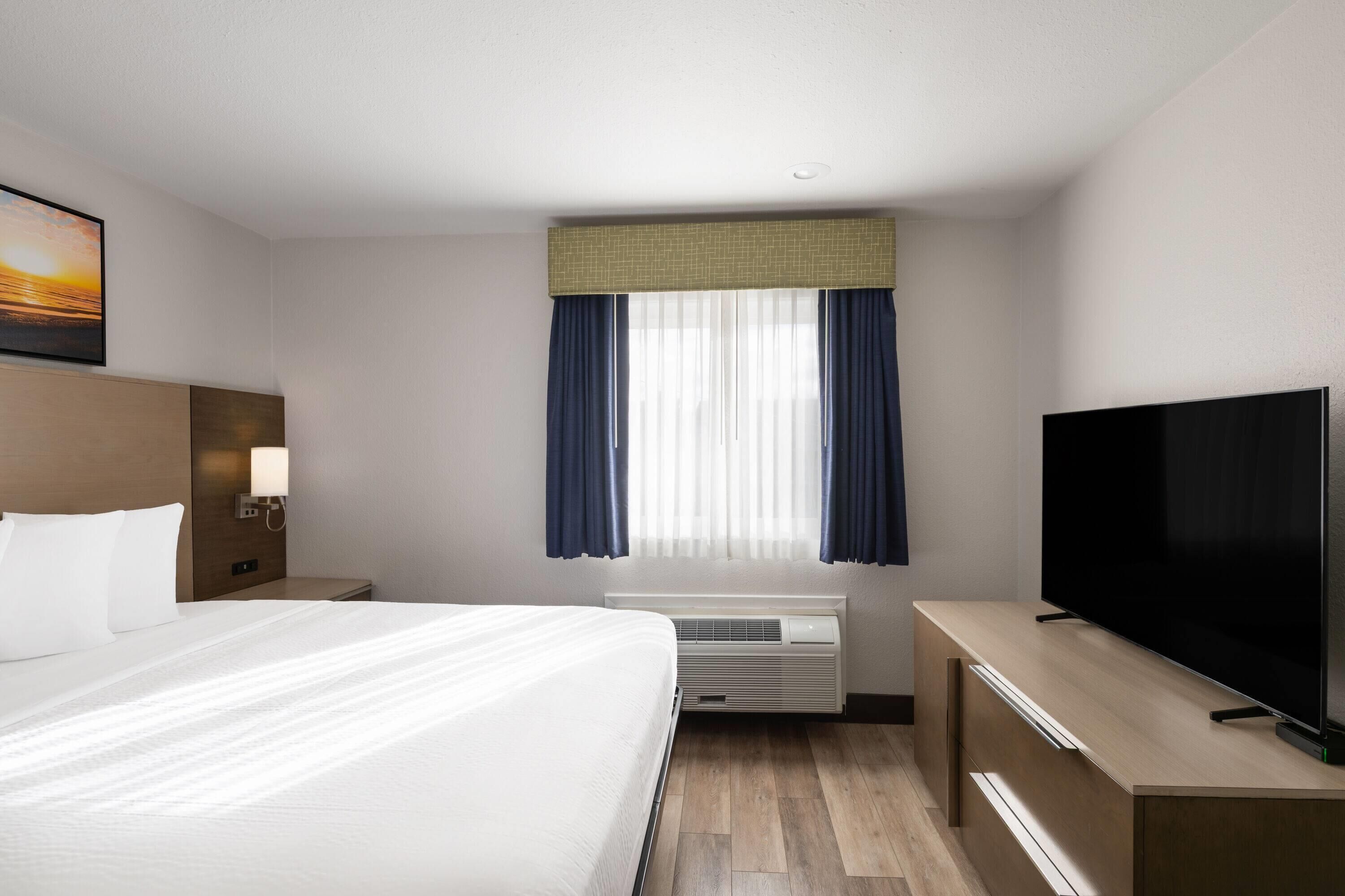 A hotel room with a bed, a TV, a desk, a nightstand, a window with curtains, and a wall-mounted picture.