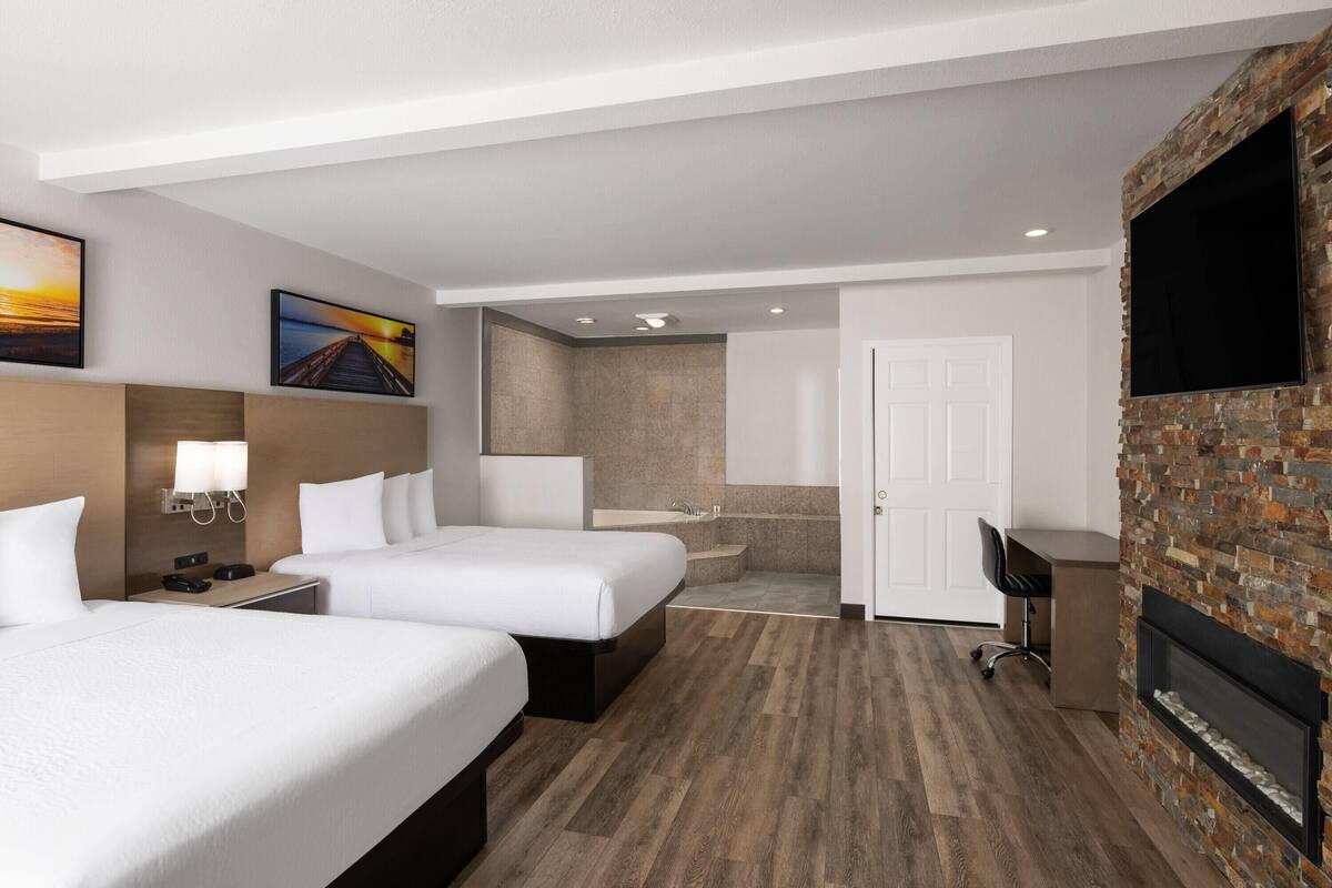 A hotel room with two beds, a desk, a TV, a fireplace, and a bathroom with a shower and sink.