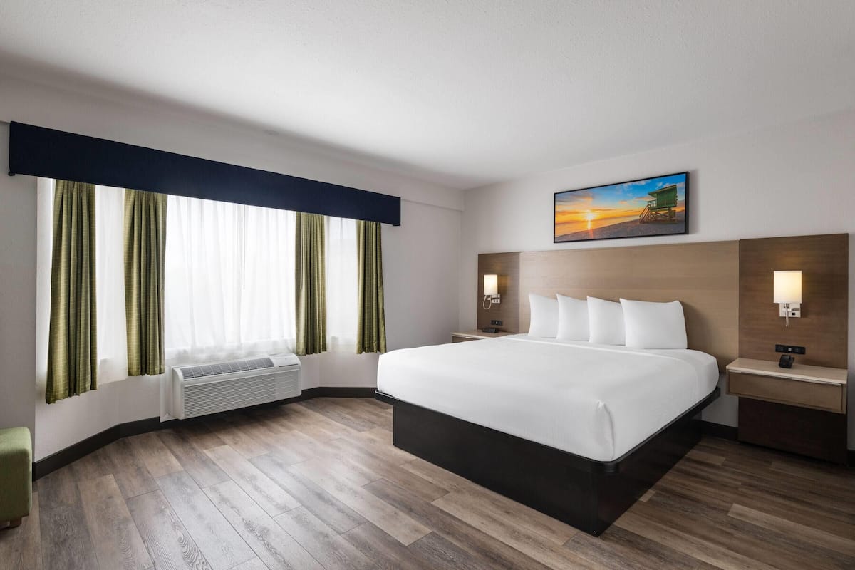 A hotel room with a large bed, a nightstand, a wall-mounted lamp, a painting of a sunset, and a window with curtains.