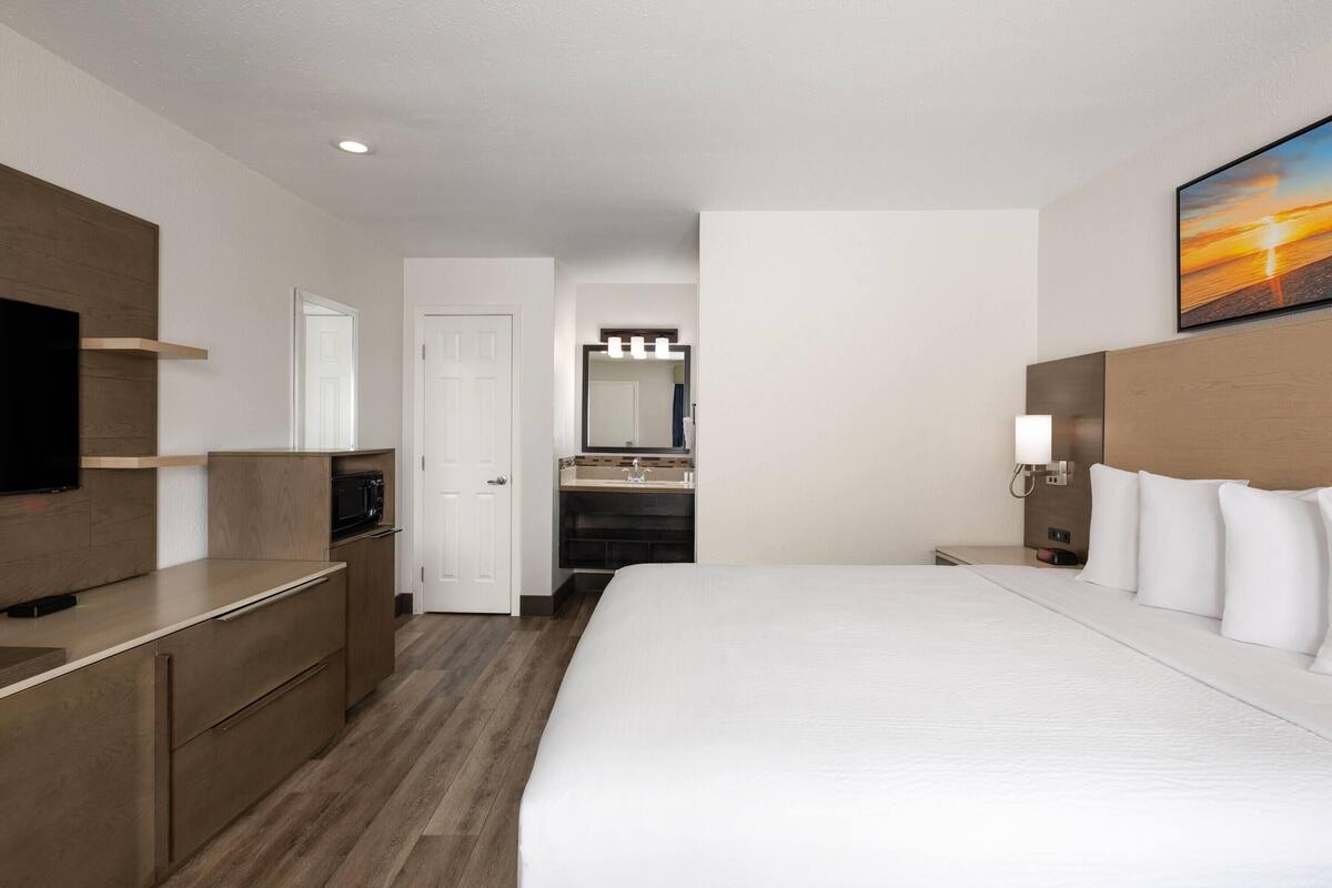A hotel room with a large bed, a TV, a wooden dresser, a bathroom with a sink and mirror, and a painting on the wall.