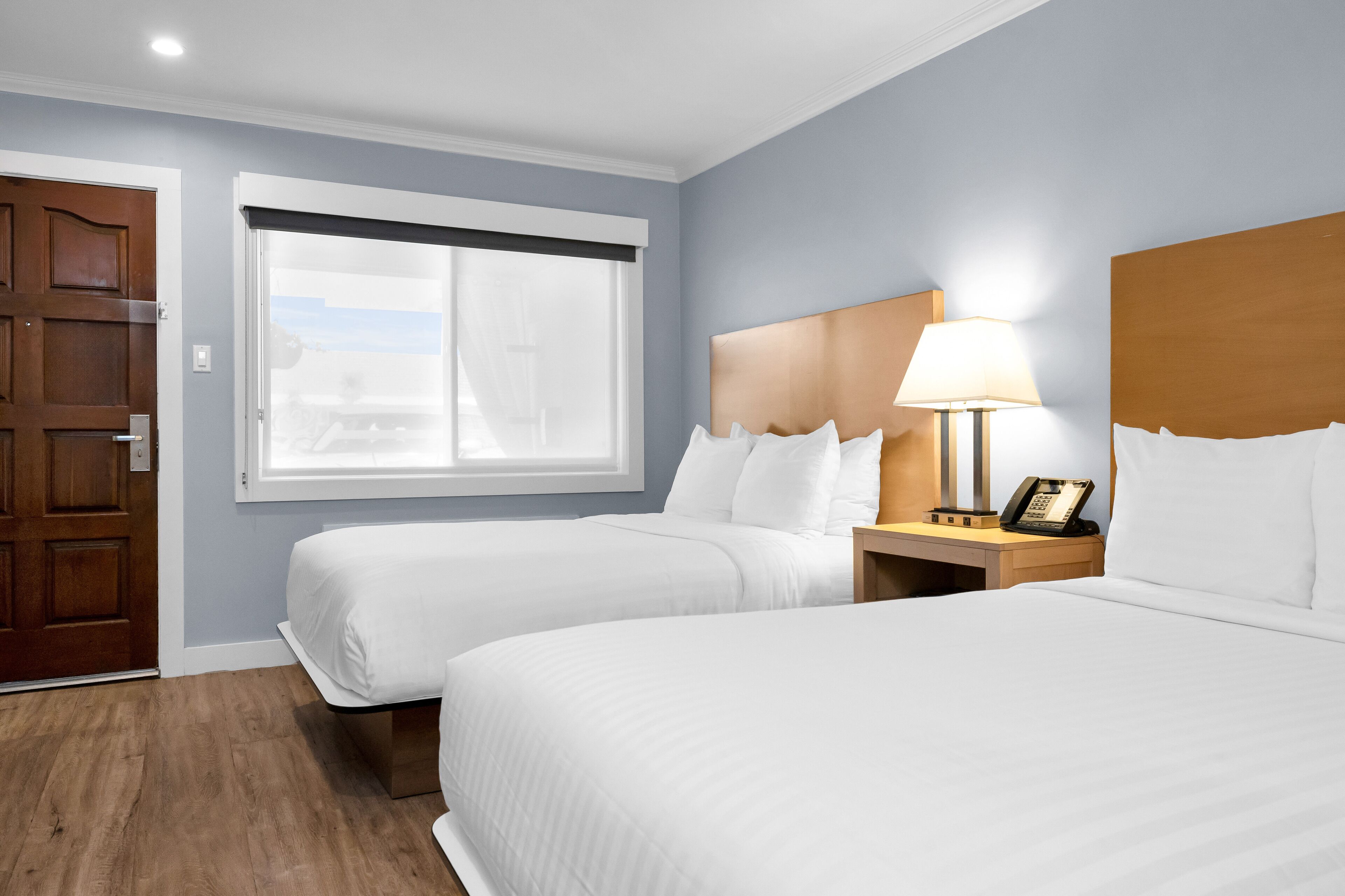 Habitación de hotel con dos camas, una puerta de madera, una ventana con persianas, una mesita de noche con lámpara y un teléfono.