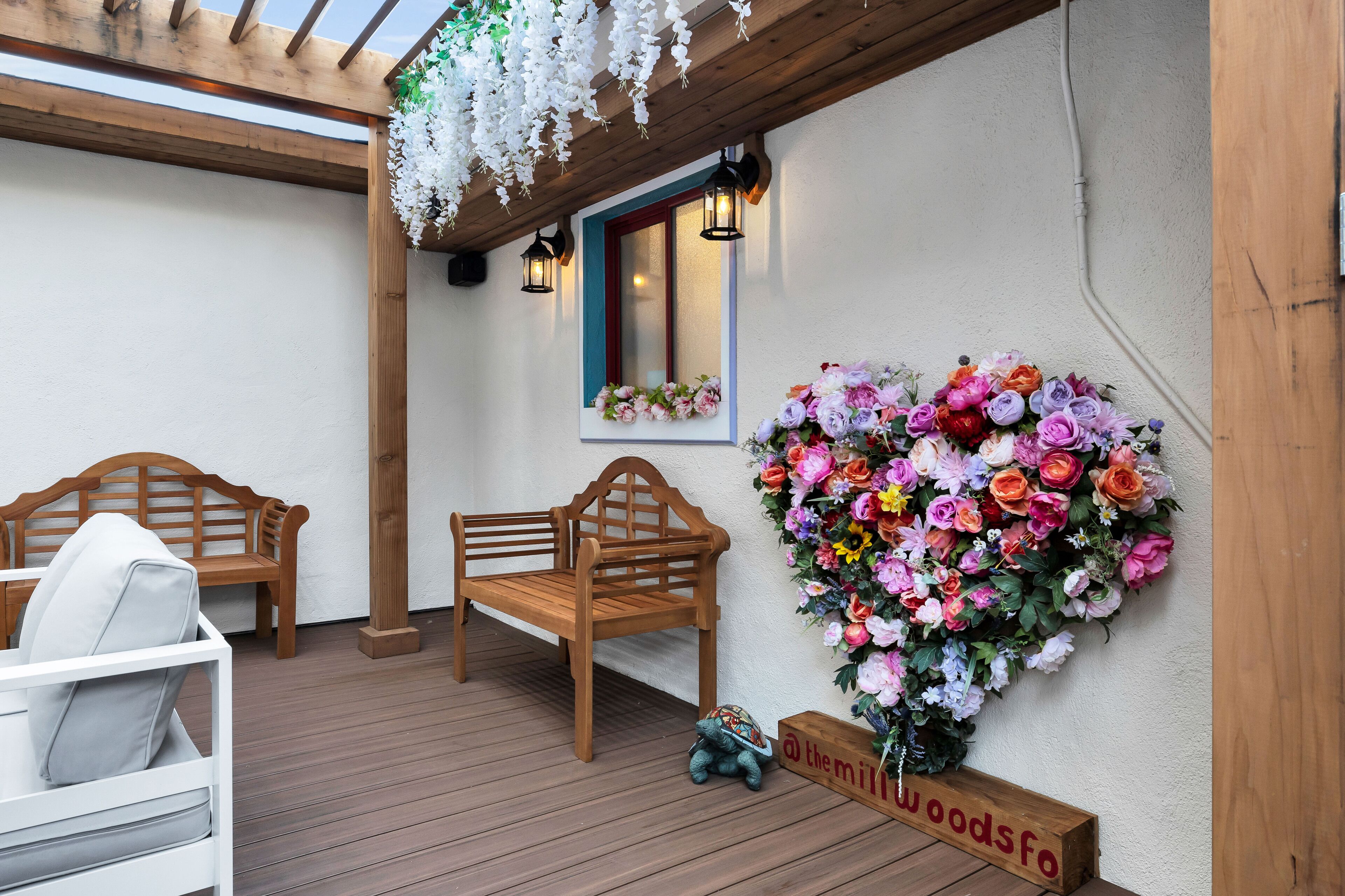 A wooden bench and a white chair under a wooden pergola with hanging white flowers.