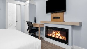 Una habitación de hotel moderna con una cama, un escritorio con una silla, un televisor de pantalla plana sobre una chimenea y una puerta que da a un baño.