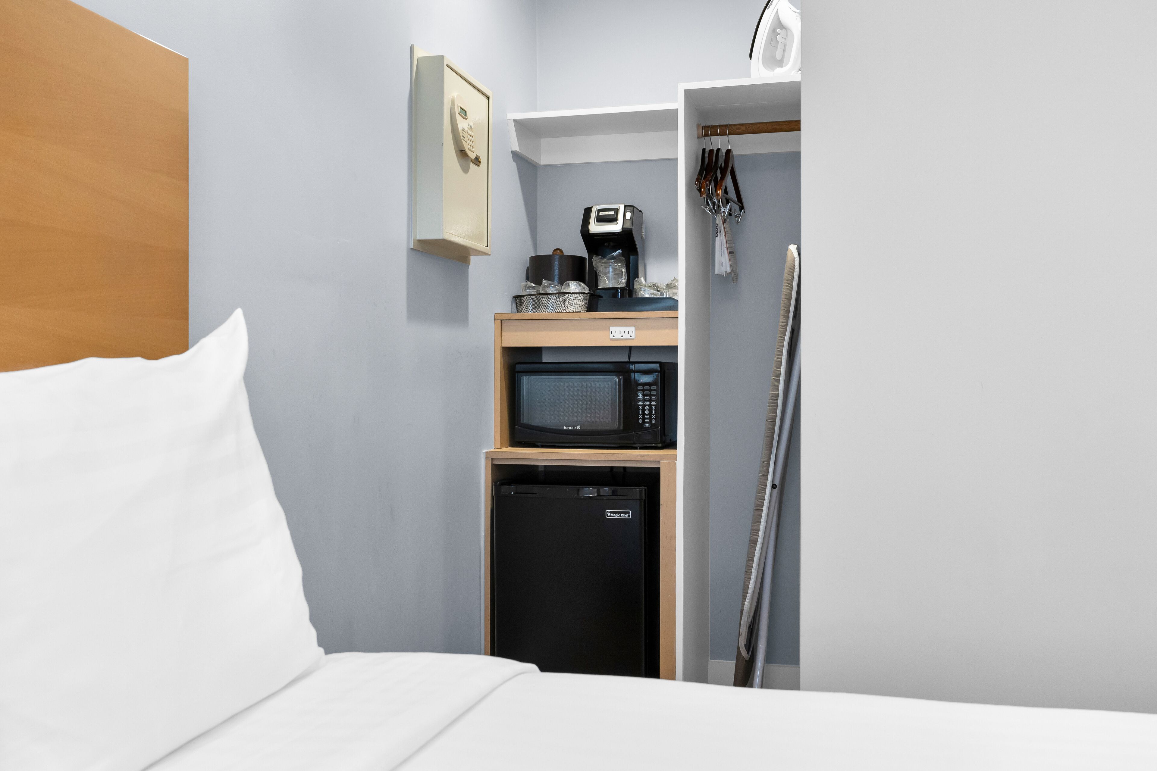 Une chambre d’hôtel compacte comprenant un lit, un four à micro-ondes, un petit réfrigérateur, une machine à café et un ensemble d’ustensiles accrochés au mur.
