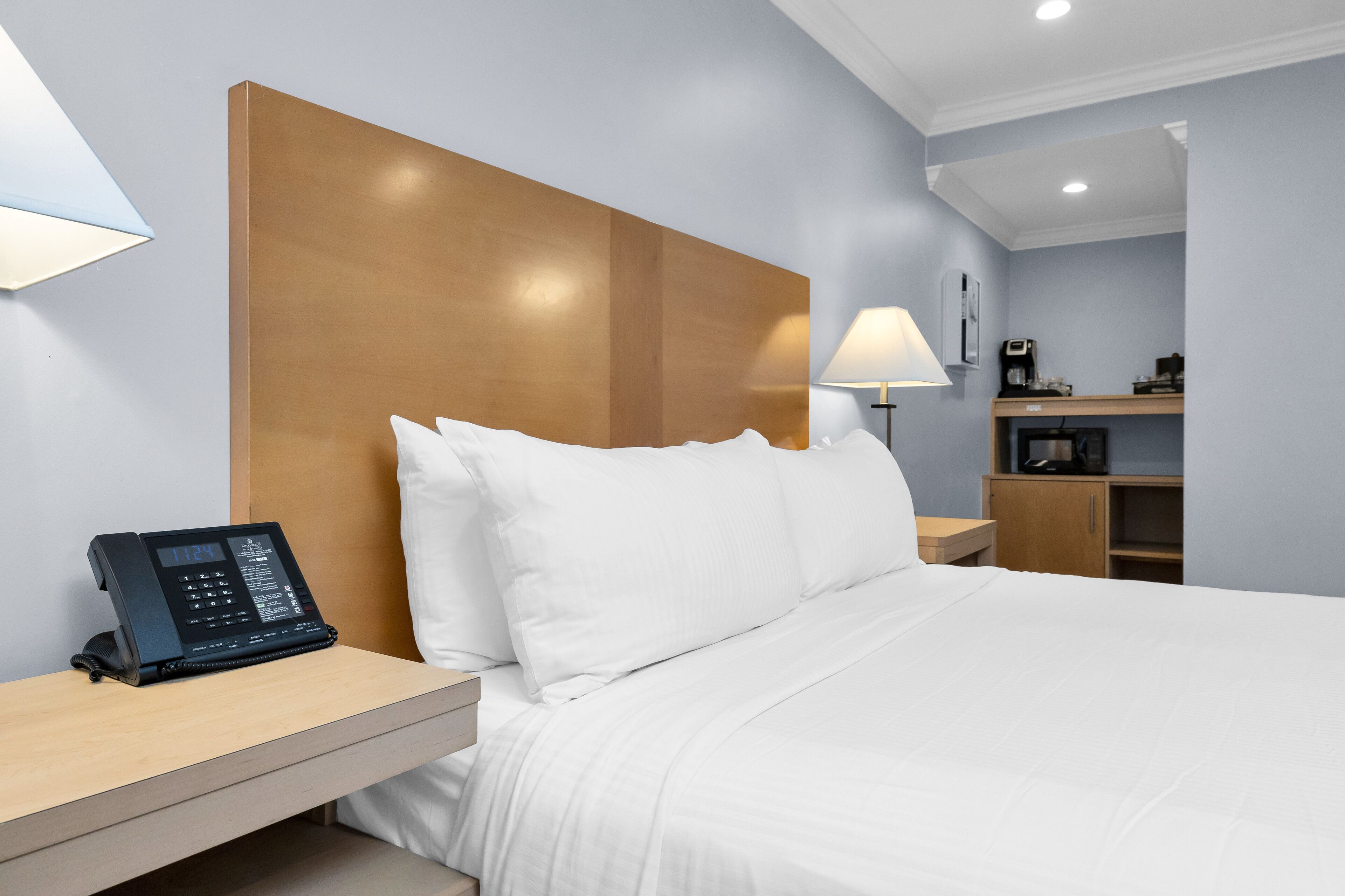 A hotel room with a wooden headboard, a bedside phone, a lamp, and a small desk with a coffee machine.