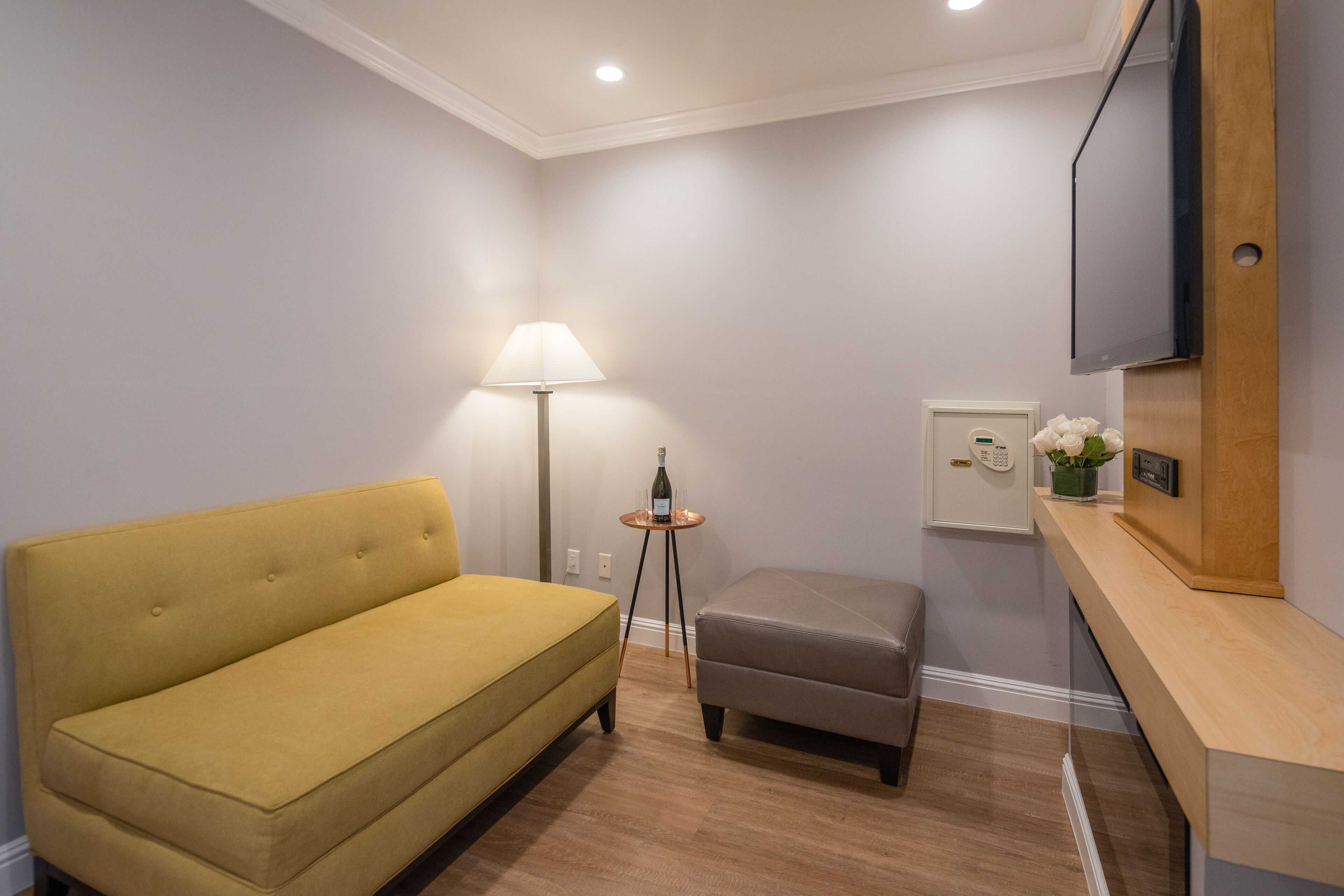 Une pièce avec un canapé jaune, un pouf gris, une petite table avec une lampe, un téléviseur fixé au mur et un comptoir en bois.