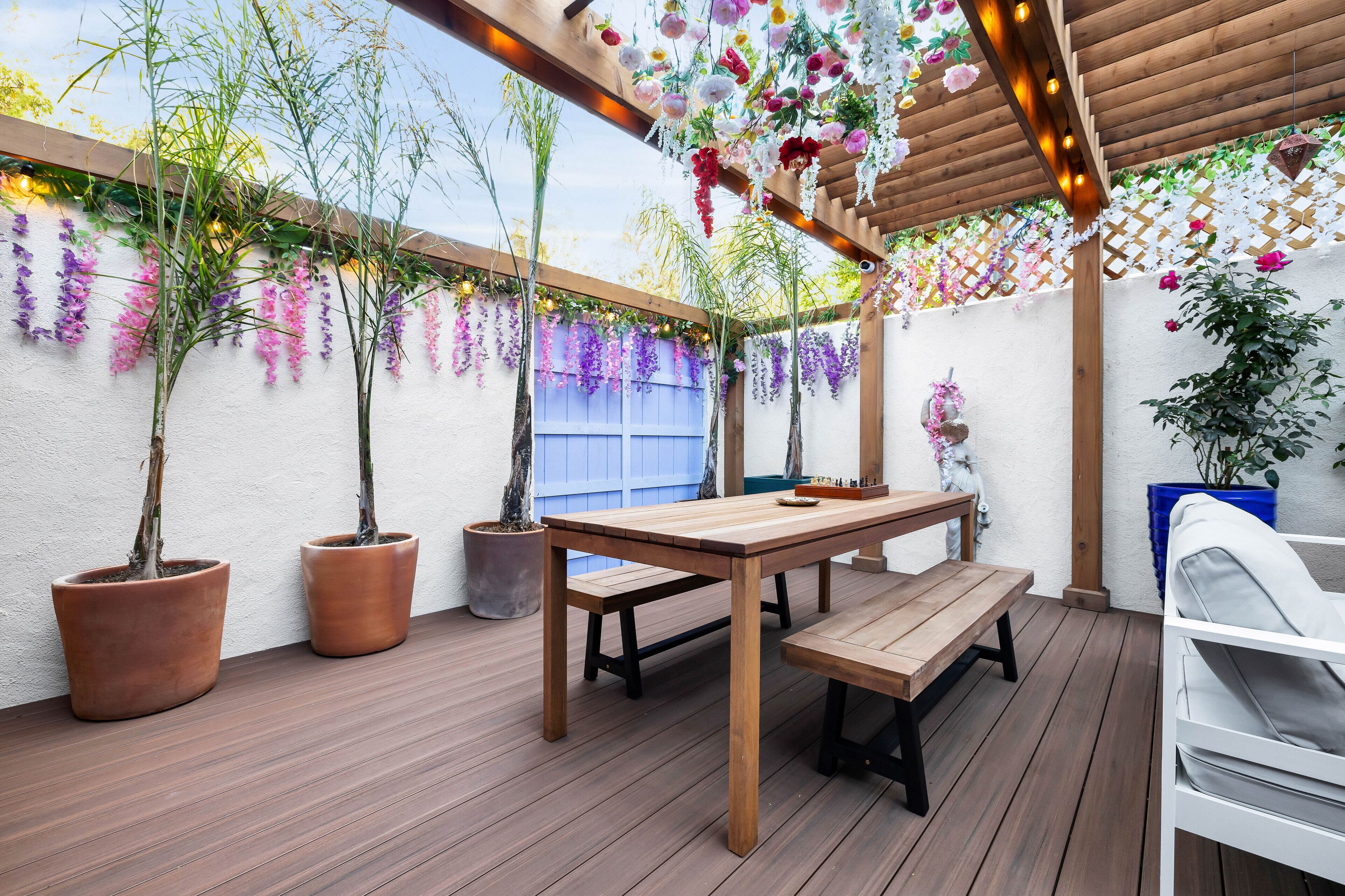 A wooden deck with a table and benches, surrounded by potted plants and hanging flowers.