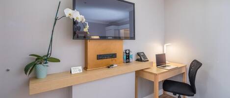 A hotel room with a wooden desk, a black office chair, a flat-screen TV mounted on the wall, a small potted plant, and a sign on the desk.