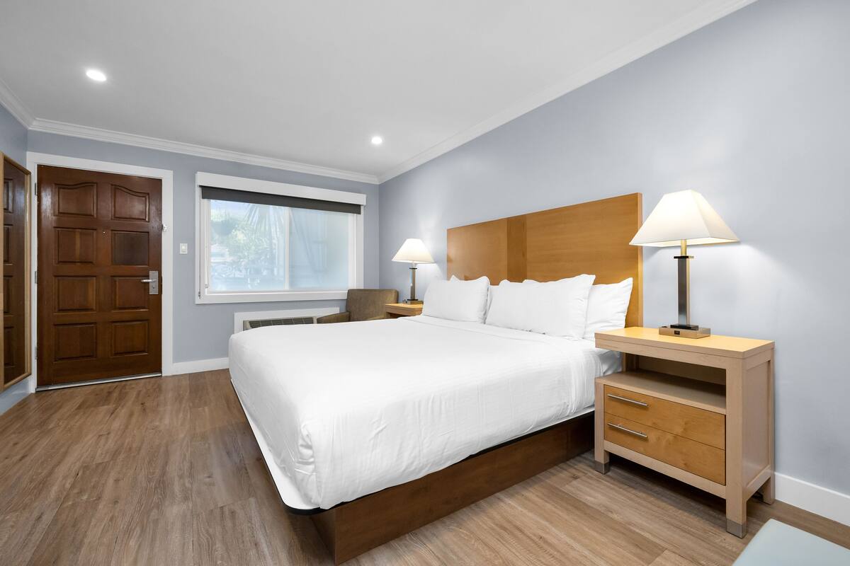 Un dormitorio moderno con una cama grande, cabecera de madera, dos mesitas de noche con lámparas, una puerta de madera y una ventana con persianas.