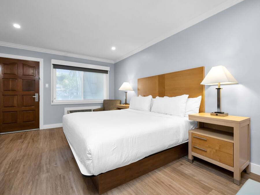 A modern bedroom with a large bed, wooden headboard, two bedside tables with lamps, a wooden door, and a window with blinds.