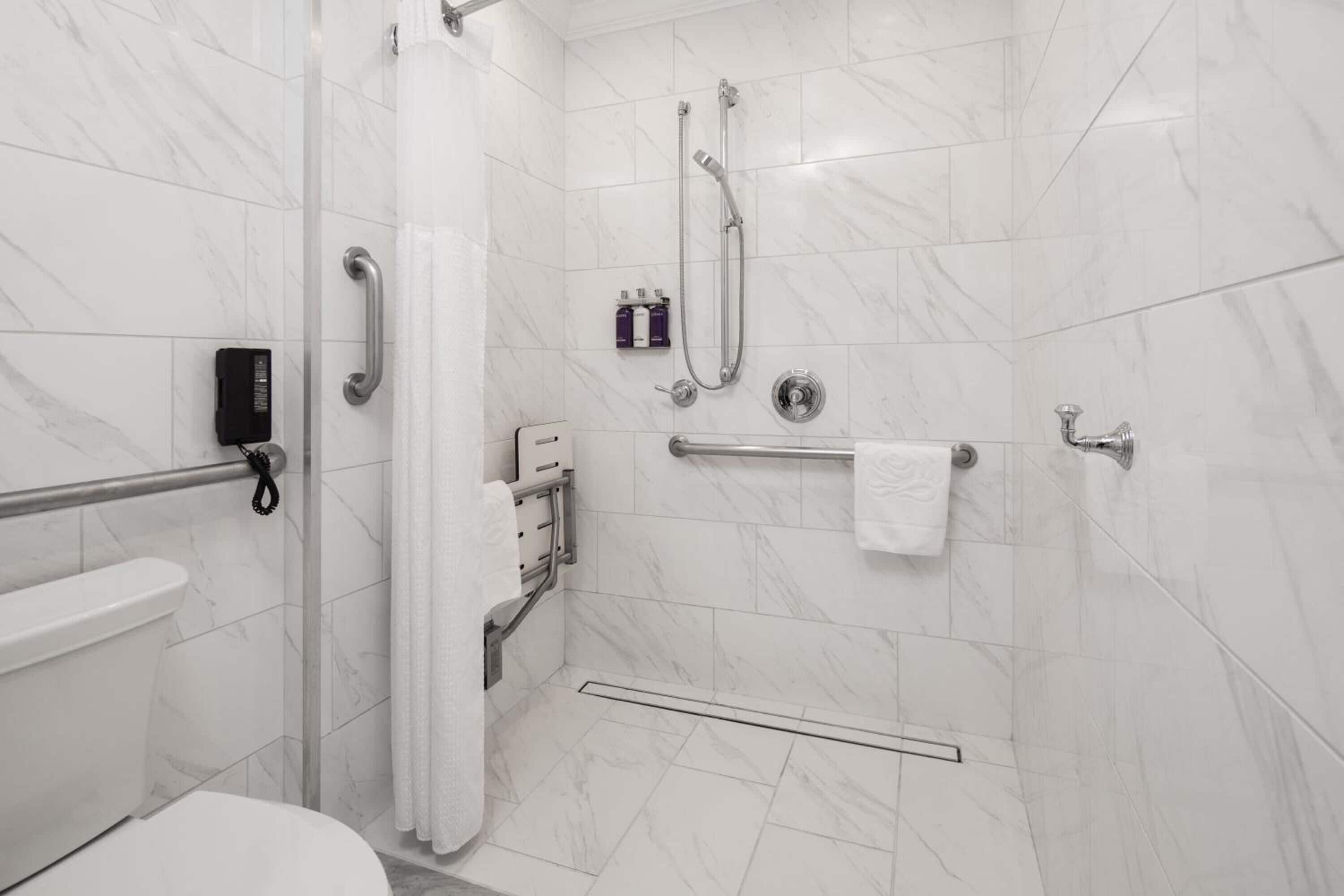 Une salle de bain moderne avec des carreaux de marbre blanc, des toilettes, une douche équipée de barres d’appui et un porte-serviettes.