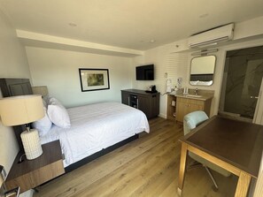A hotel room with a bed, bedside table, lamp, TV, desk, chair, and a bathroom with a mirror and sink. - Lamplighter Inn & Suites (San Luis Obispo)