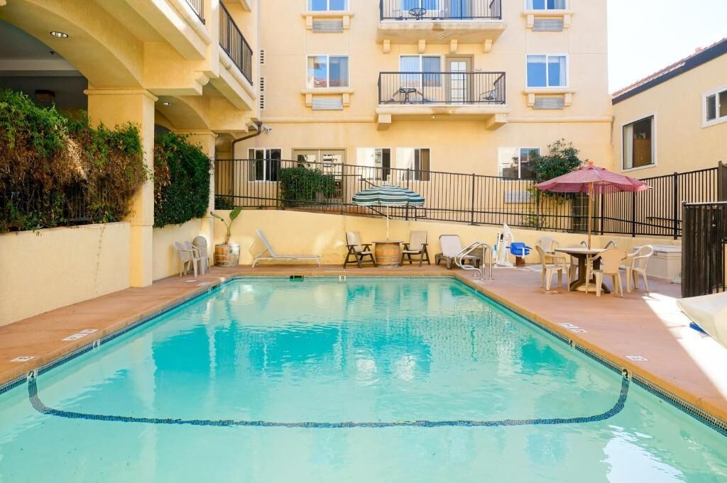 Ein Pool mit Liegestühlen und Sonnenschirmen, umgeben von einem Gebäude mit Balkonen und einem Zaun.