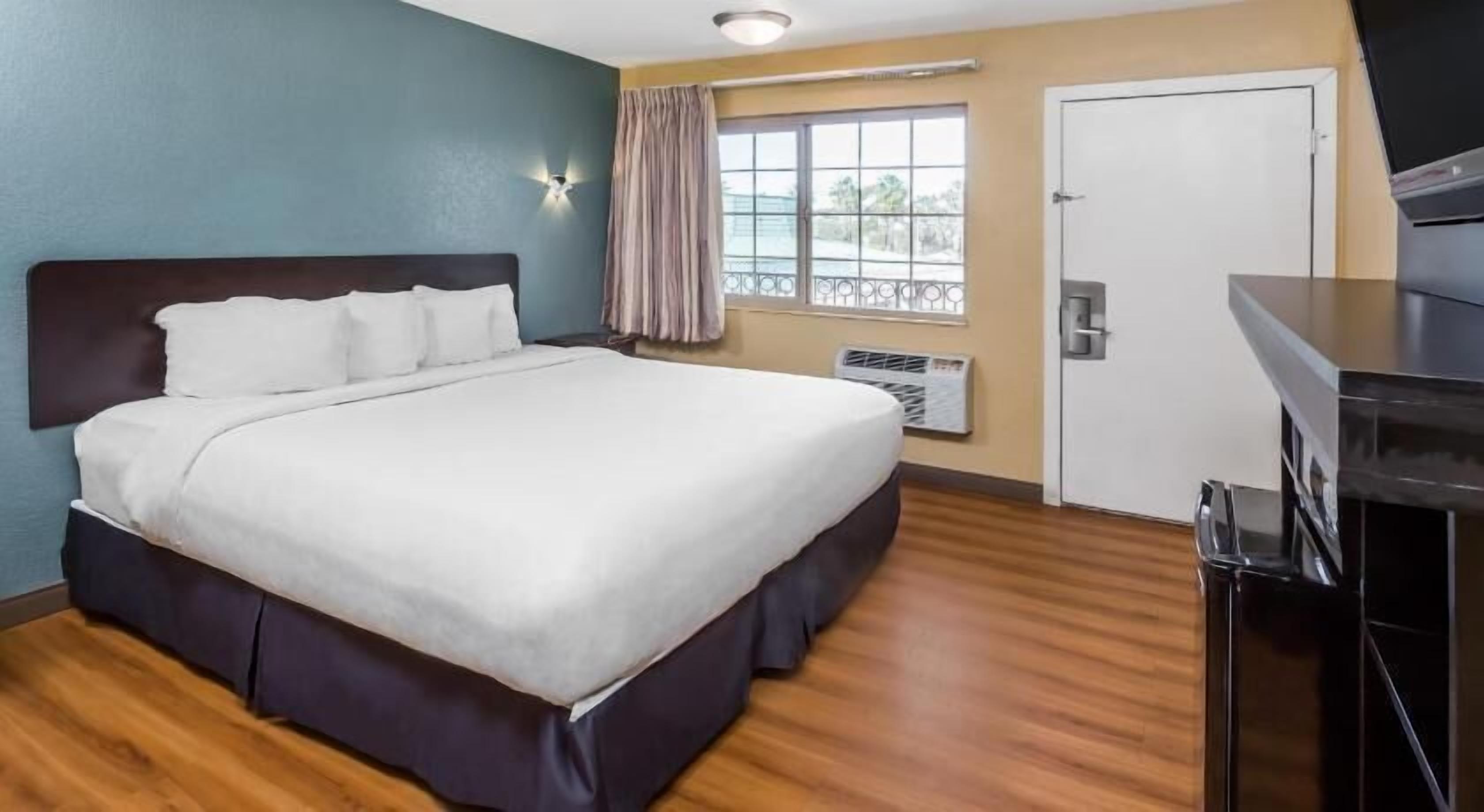 A hotel room with a large bed, a desk, a window with curtains, and an air conditioning unit.