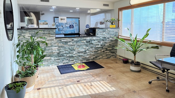 A reception area with a stone wall, a reception desk, a large window with blinds, and several potted plants. - Super 8 by Wyndham West Fargo Main Ave ND (West Fargo)