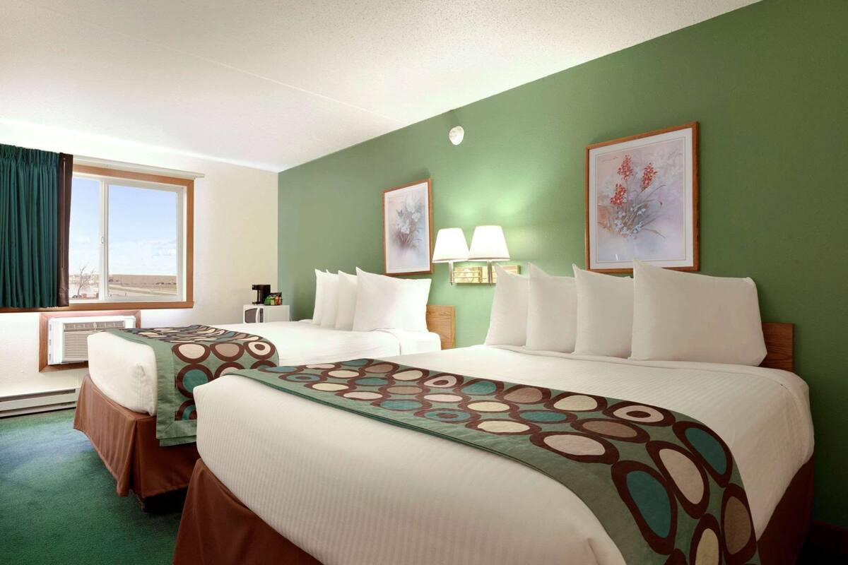 A hotel room with two beds, a desk, a chair, a window with curtains, and two wall-mounted lamps.