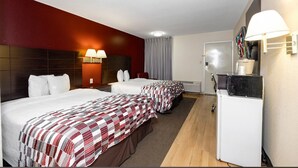 A hotel room with two beds, a desk, a refrigerator, and a wall-mounted lamp.
