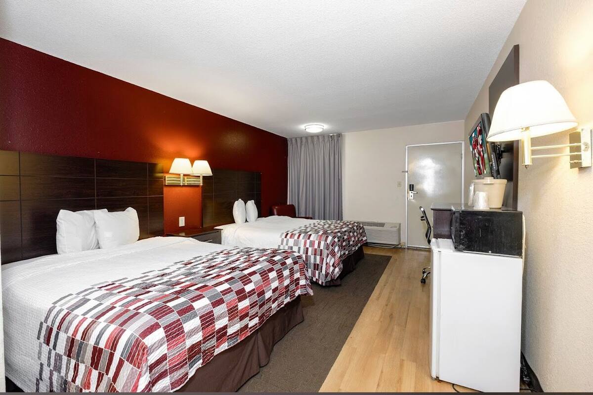 Habitación de hotel con dos camas, un escritorio, un refrigerador y una lámpara de pared.