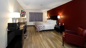 A hotel room with a bed, a desk with a computer, a red leather chair, and a TV on the wall.
