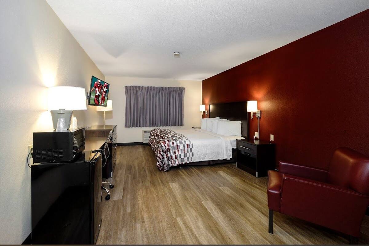 Habitación de hotel con cama, escritorio con computadora, sillón de cuero rojo y televisor en la pared.