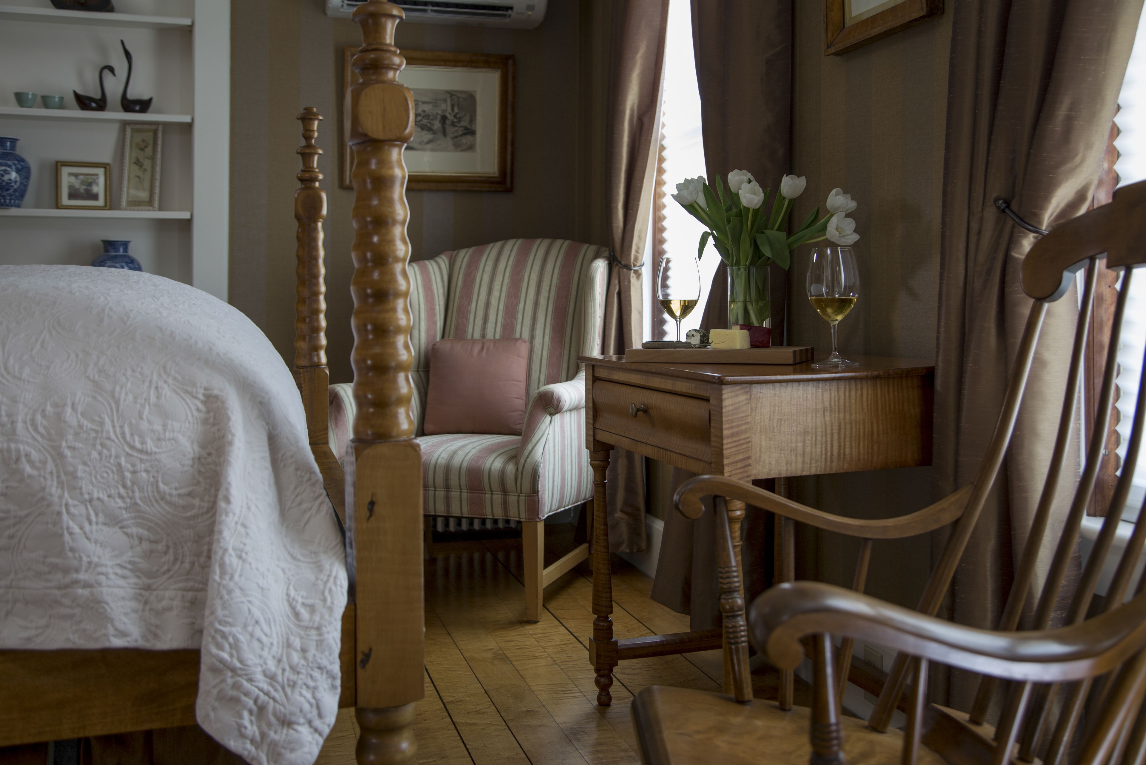 A bedroom with a bed, a wooden chair, a desk with a vase of flowers, and a window with curtains.