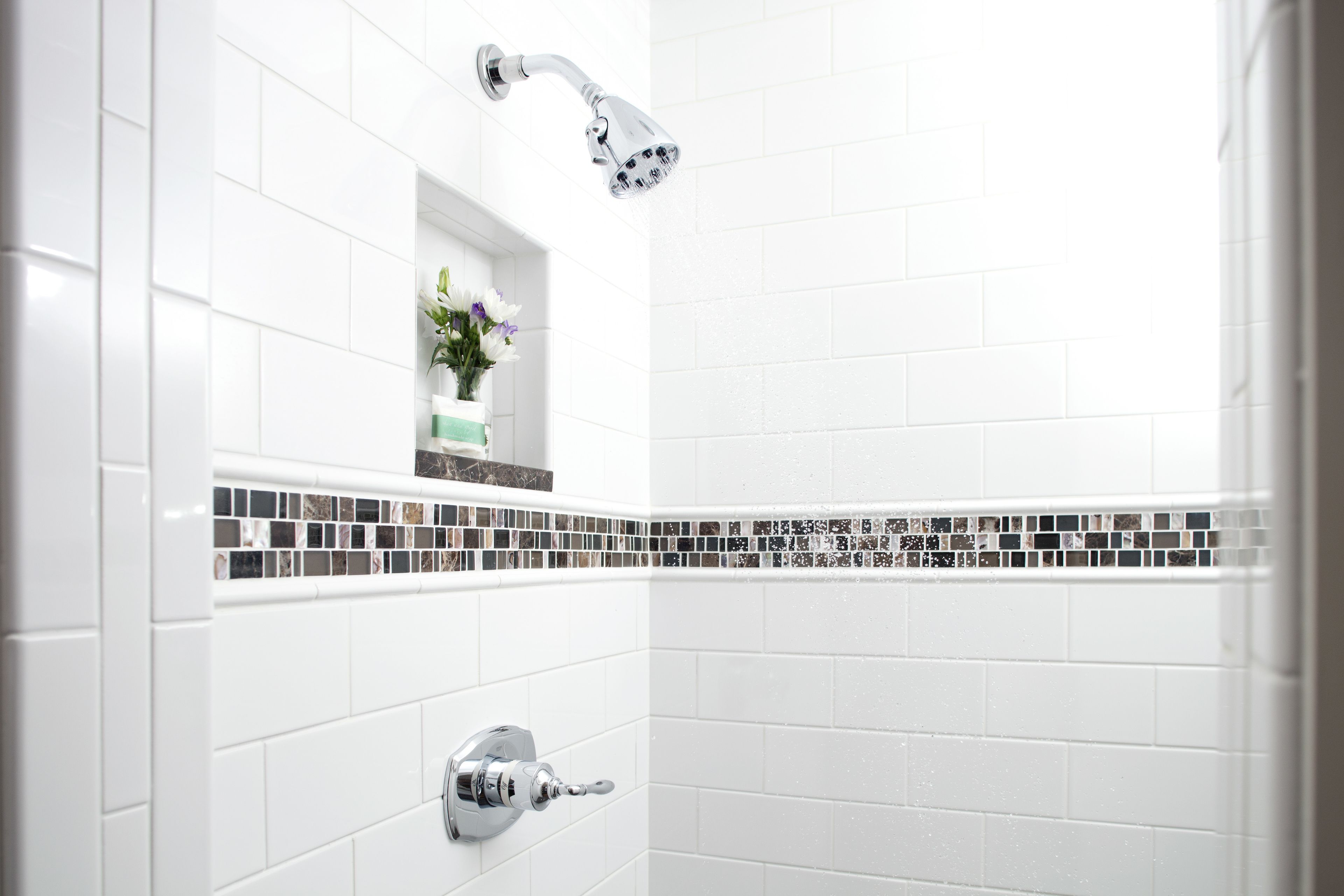 A bathroom with white tiles, a showerhead, and a small shelf with flowers.