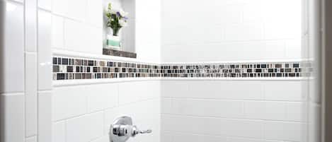 A bathroom with white tiles, a showerhead, and a small shelf with flowers.