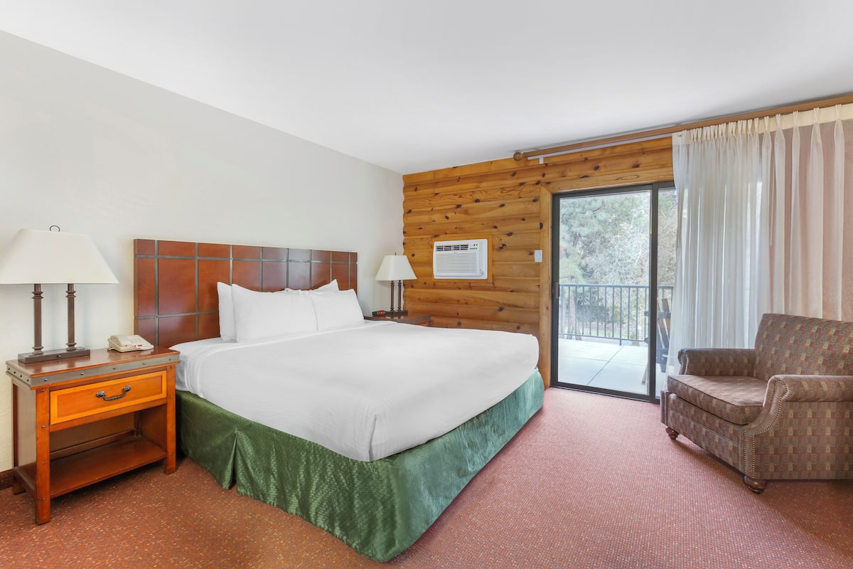 A hotel room with a wooden headboard bed, a green bed skirt, a nightstand with a lamp, a chair, and a sliding glass door to the outside.