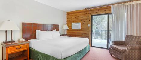 Une chambre d’hôtel avec un lit doté d’une tête de lit en bois, un couvre-lit vert, une table de chevet avec une lampe, une chaise et une porte-fenêtre coulissante donnant sur l’extérieur.