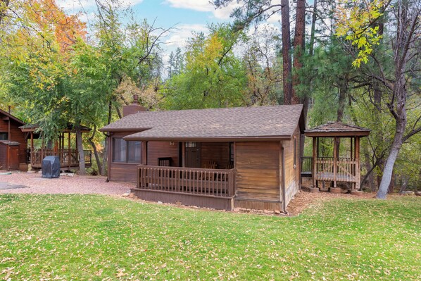 A rustic cabin with a porch, surrounded by trees and a grassy yard.