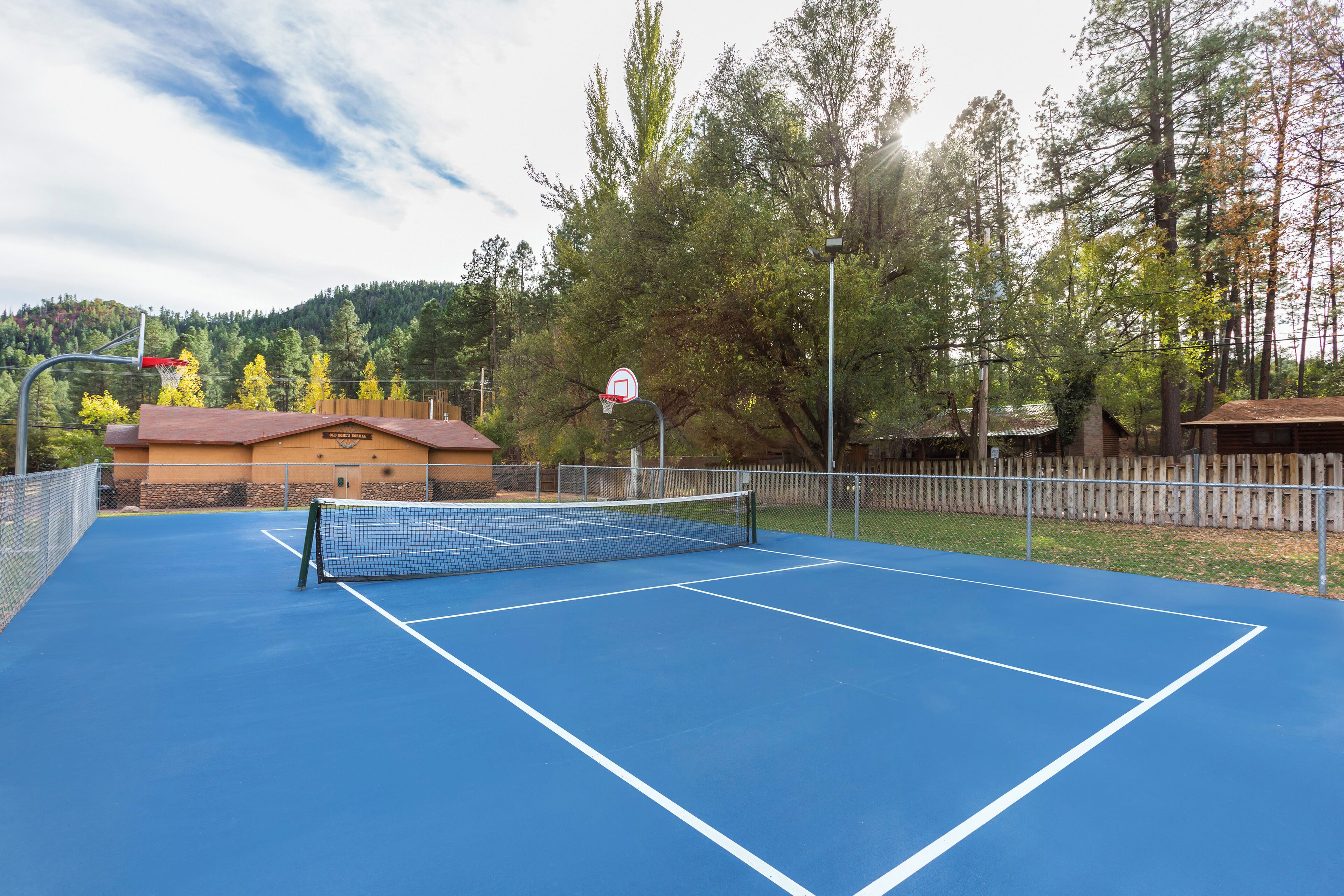 An outdoor tennis court with a net, surrounded by a fence and trees.