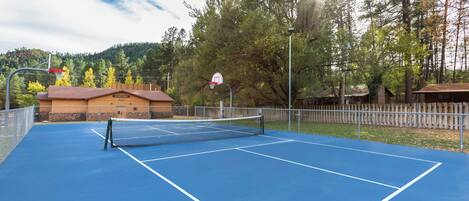 An outdoor tennis court with a net, surrounded by a fence and trees.
