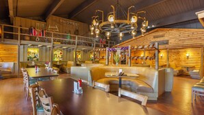 Un restaurant spacieux avec de hauts plafonds en bois, de nombreux espaces de restauration, dont une grande table centrale et des banquettes, et un escalier apparent menant à un étage supérieur.