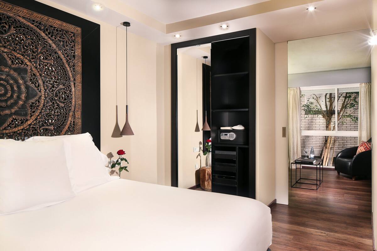 A modern hotel room with a large bed, a decorative wall hanging, a small table with a vase of flowers, and a view of the outside through a window.