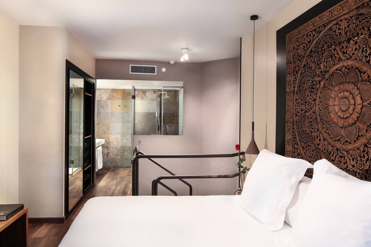 A modern hotel room with a large bed, a glass shower enclosure, and a decorative wall panel.