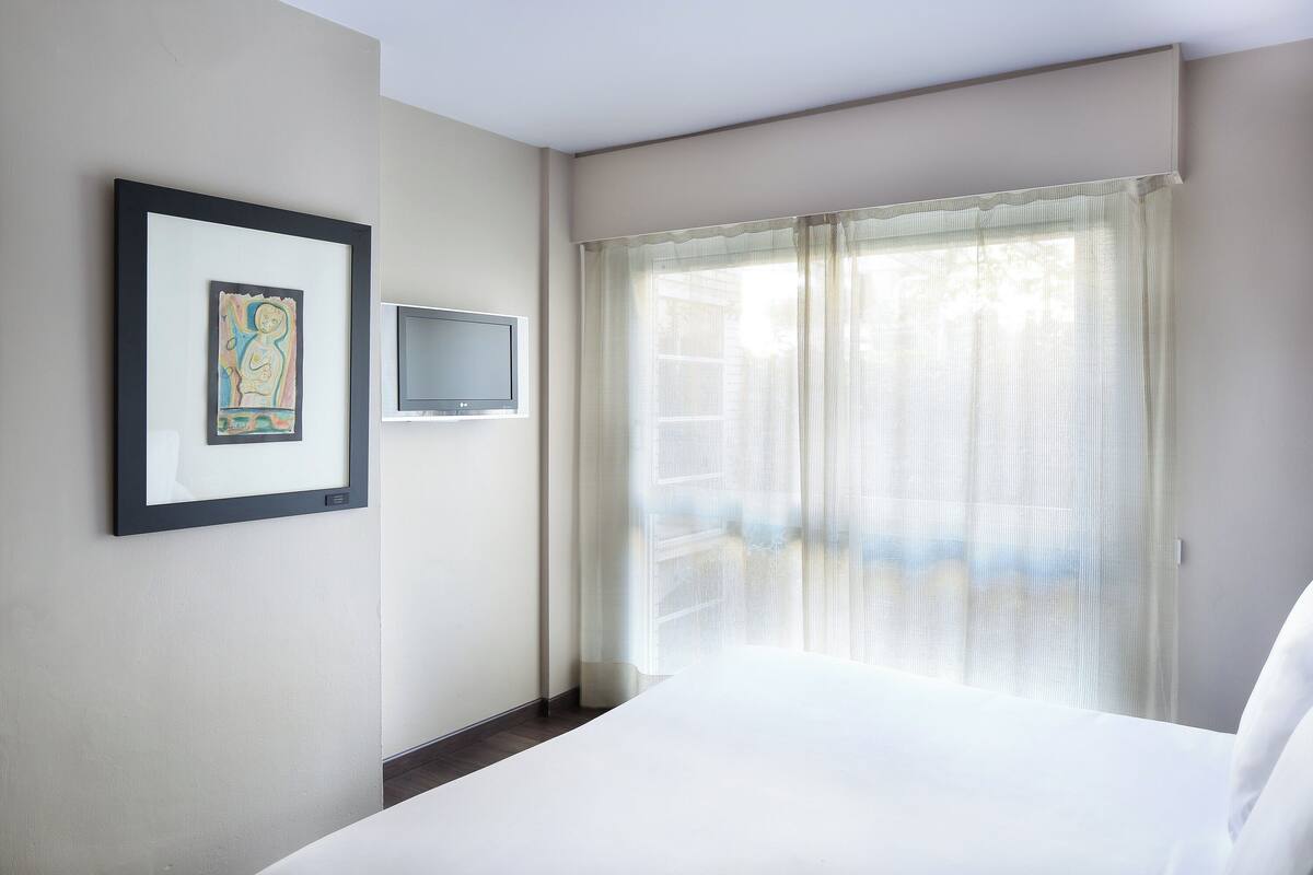 A hotel room with a bed, a television mounted on the wall, and a window with sheer curtains.