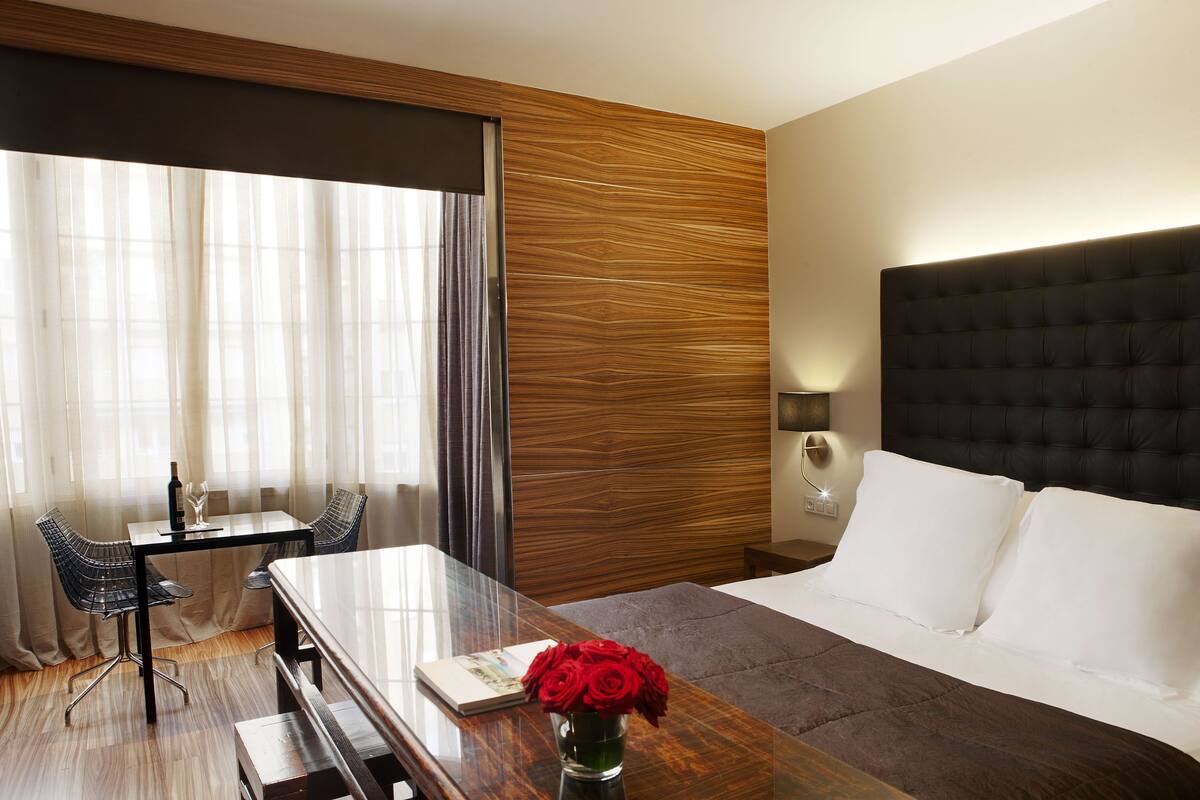 A hotel room with a large bed, a wooden headboard, a desk with a chair, a small table with a vase of red roses, and a large window with sheer curtains.
