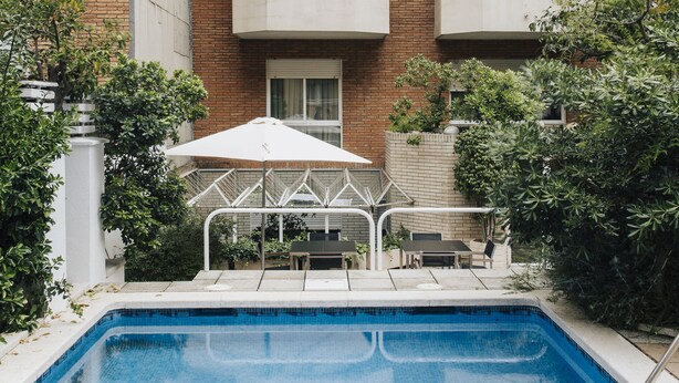 A swimming pool with a white umbrella, surrounded by greenery and a brick building with multiple windows. - Gran Derby Suites Hotel, a Small Luxury Hotels of the World (Barcelona)