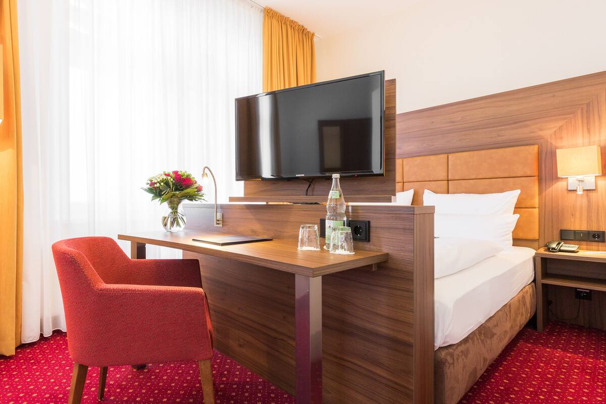 A hotel room with a bed, a desk with a chair, a TV, a bedside table with a lamp, and a red carpet.