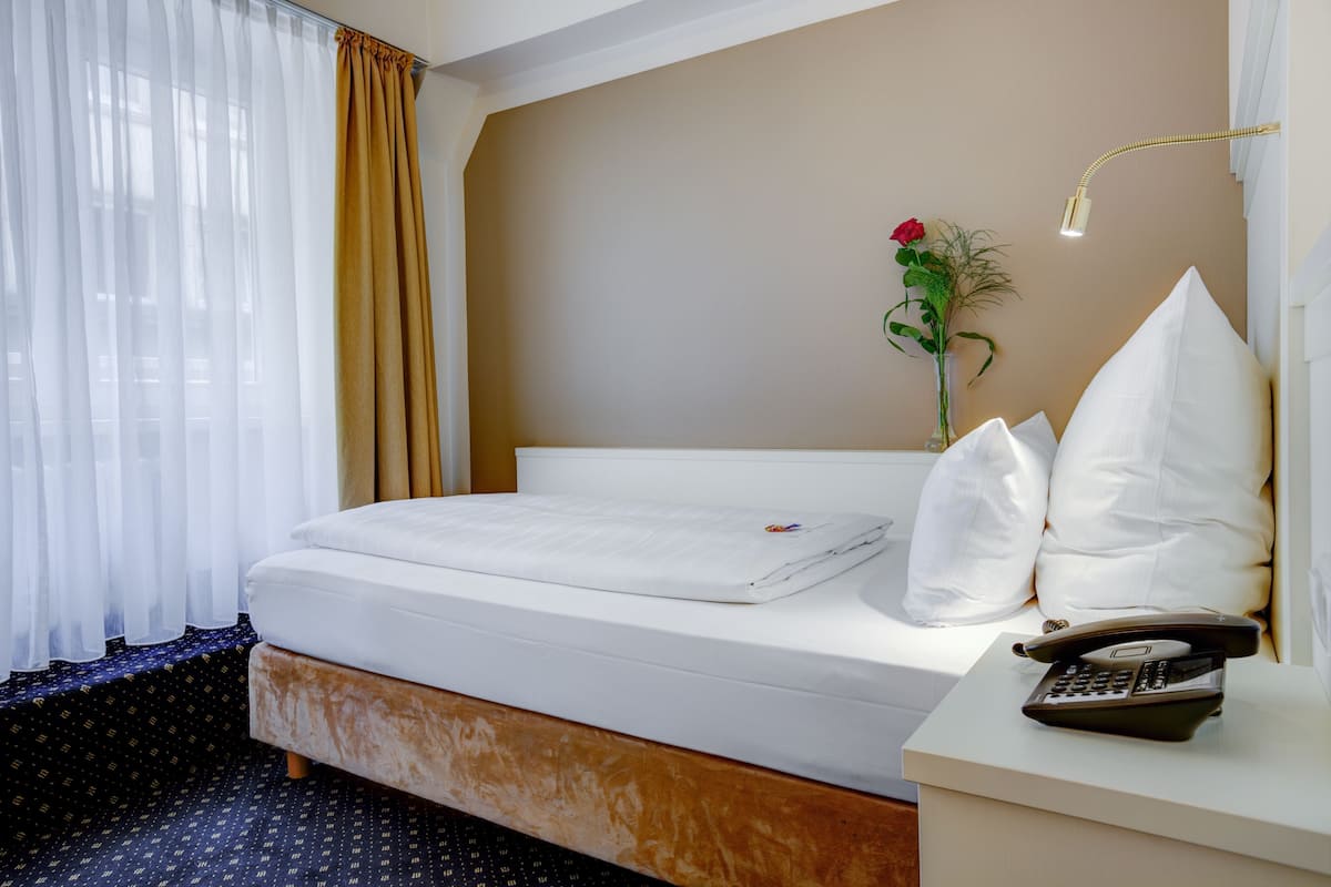 A hotel room with a bed, a bedside table with a telephone, a vase with a flower, and a window with curtains.