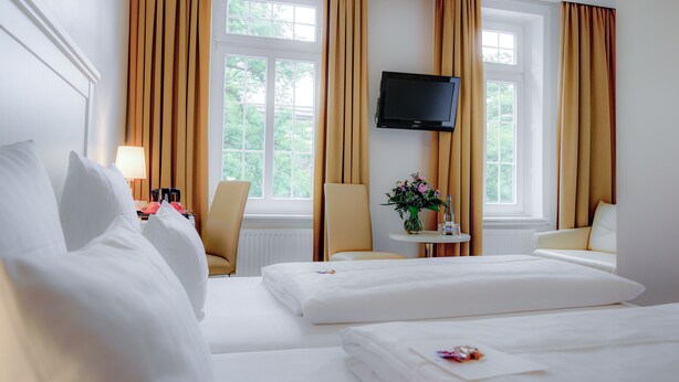 Habitación de hotel con dos camas, un televisor montado en la pared, una silla, una mesita con un jarrón de flores y una ventana con cortinas.