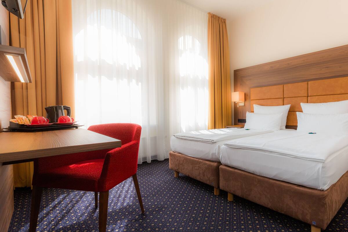 A hotel room with two beds, a desk with a coffee maker, a red chair, and a nightstand with a phone.