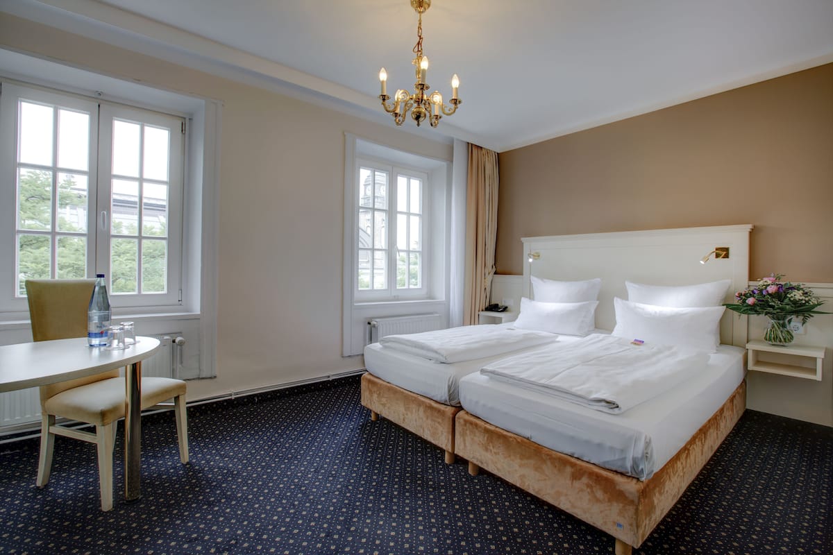 A hotel room with a double bed, a small table with a chair, a desk, and a window with curtains.