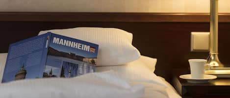 A neatly made bed with a book titled "MANNHEIM" on it, a white cup on a saucer, and a lamp on a bedside table.