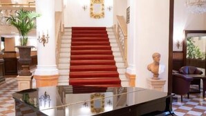 Un magnifique piano à queue trône dans un hall spacieux, dont le sol est recouvert d’un motif à carreaux. Un escalier à moquette rouge et un lustre complètent le décor.