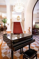 Um piano de cauda num hall espaçoso com chão quadriculado, uma escadaria com carpete vermelho e um lustre.