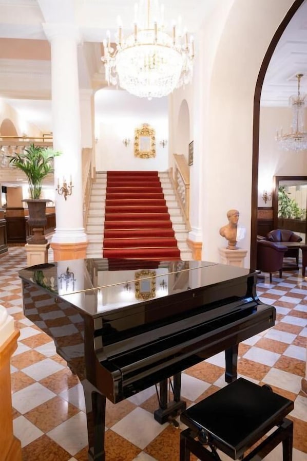 Un piano de cola en un vestíbulo amplio con piso a cuadros, una escalera con alfombra roja y un candelabro.