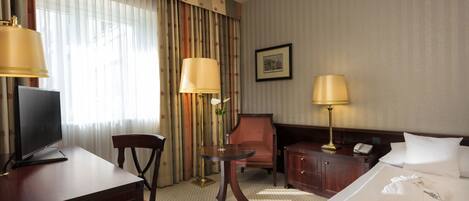 A hotel room with a bed, desk, chair, lamps, and a framed picture on the wall.