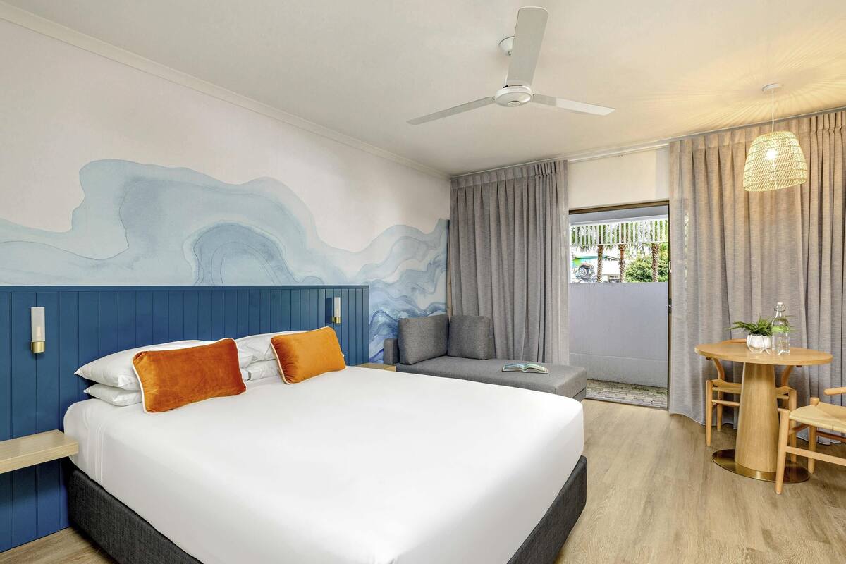 A modern hotel room with a large bed, a seating area, a dining table, and a ceiling fan.