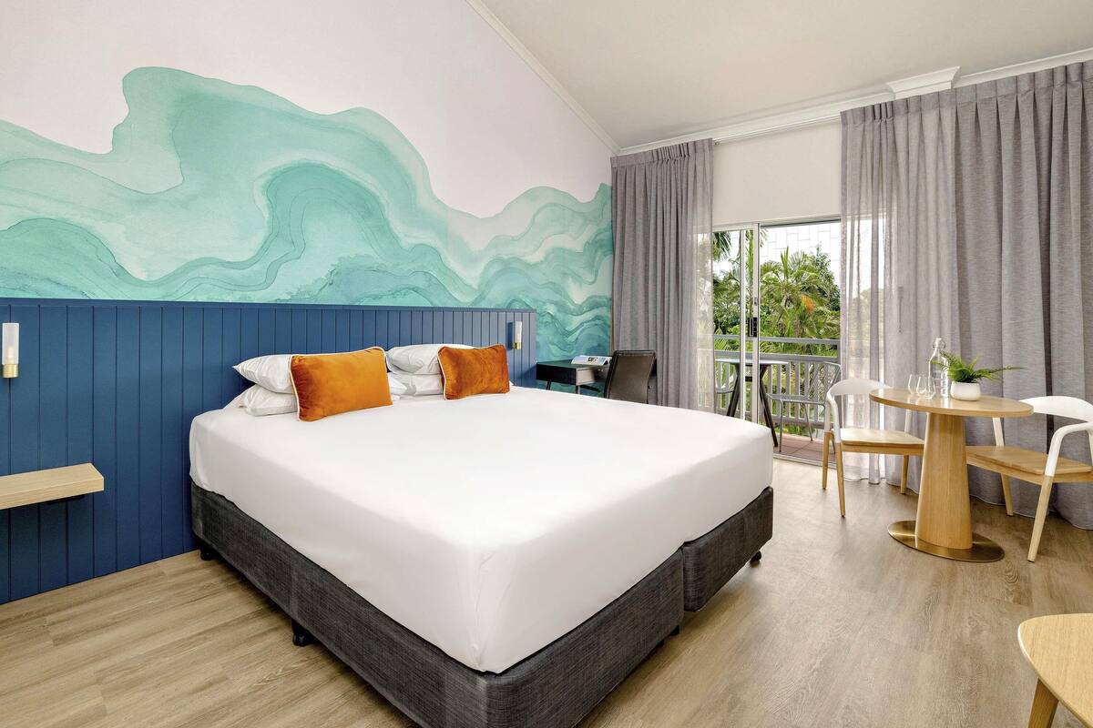A hotel room with a large bed, a desk with a chair, a balcony with a table and chairs, and a view of greenery outside.